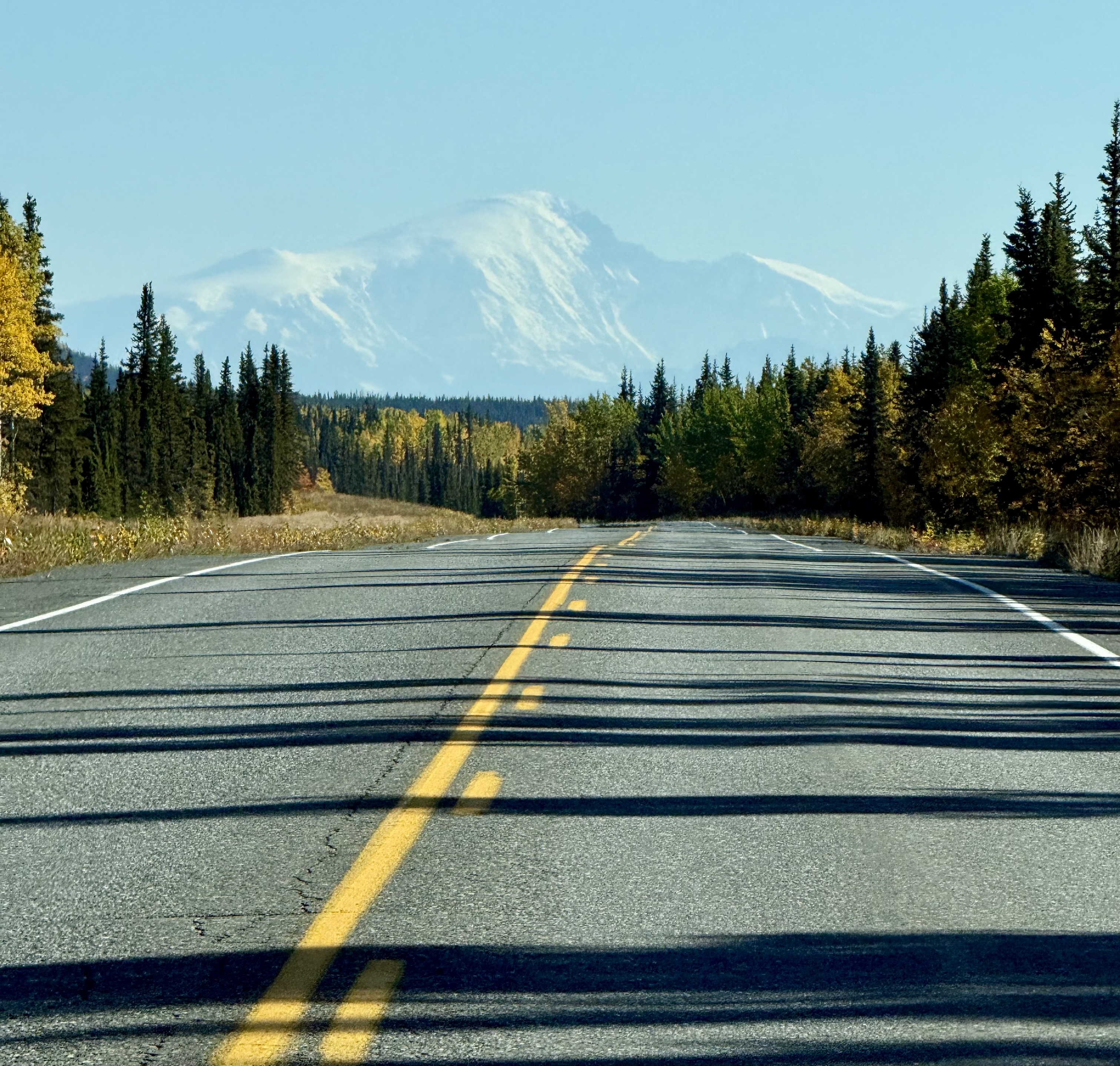 “Highway bordered by autumn trees leading toward a snow-covered mountain beneath a blue sky.”